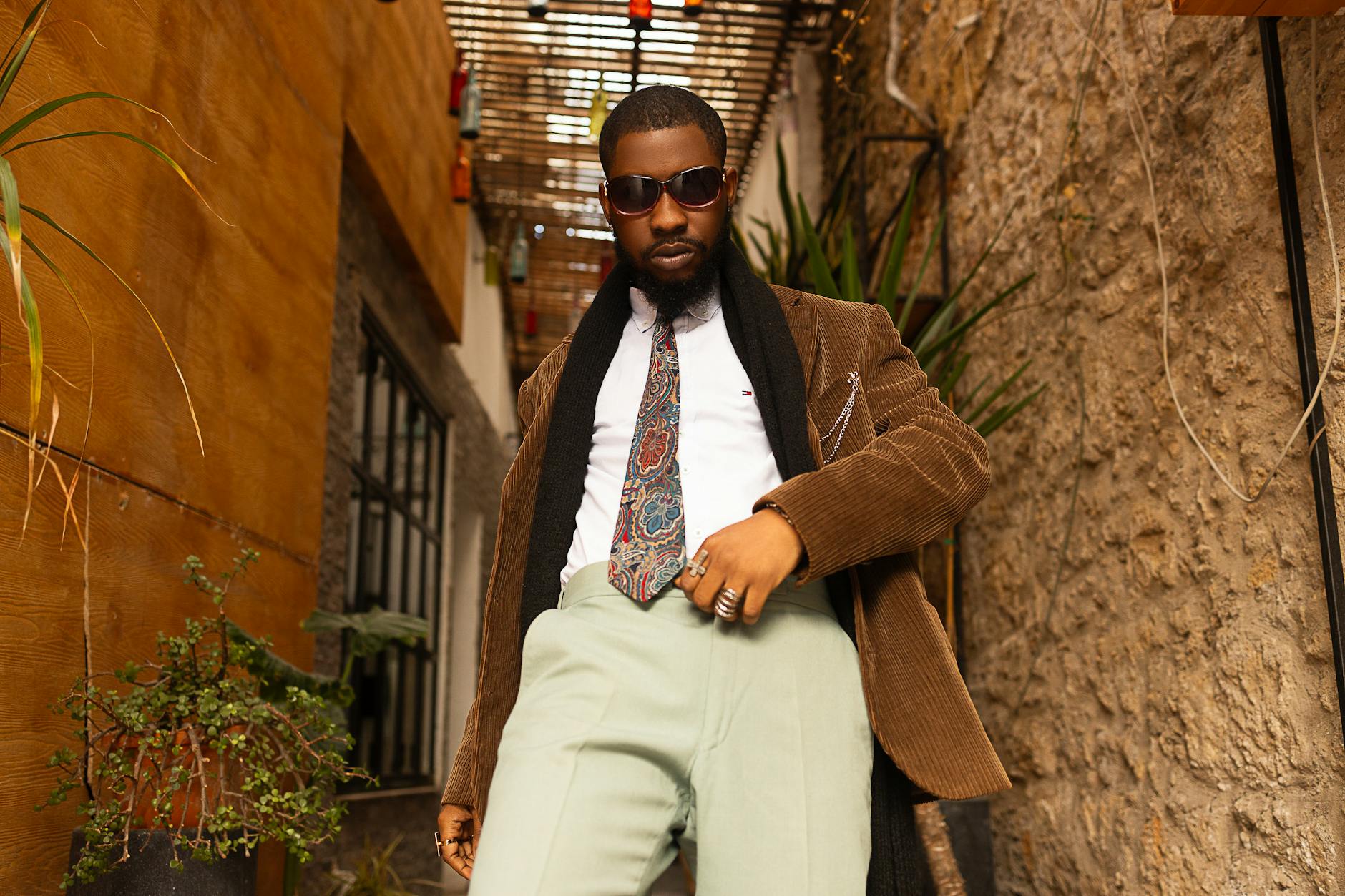 A fashionable man in a corduroy jacket poses confidently in an urban setting, wearing sunglasses and a paisley tie.